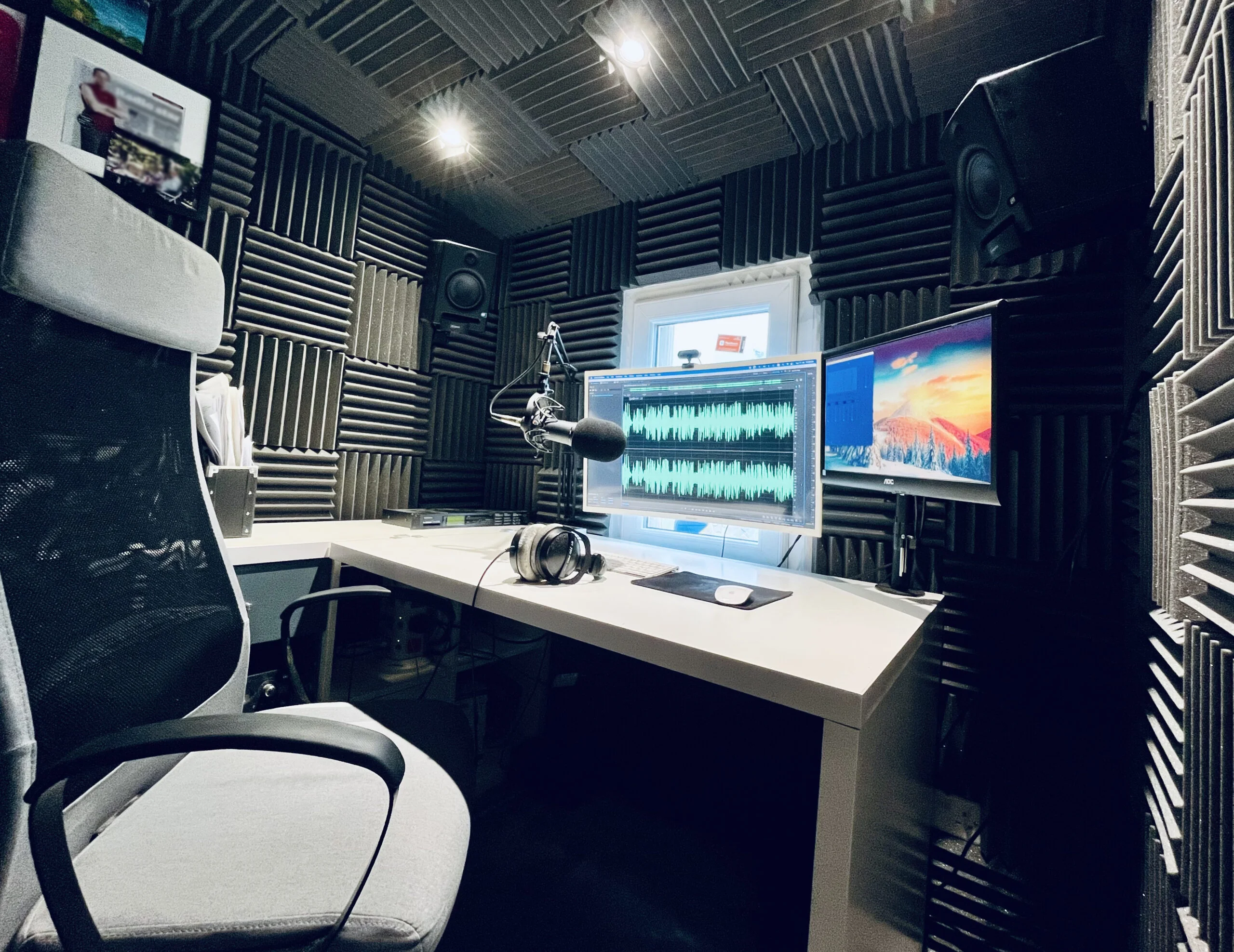 Image of the home studio Roberto uses for his voice tracking presenter services. To the left of the screen is his big chair, in the middle is the white desk which has two computer monitors above it, along with his microphone. The walls are all covered in acoustic foam for sound proofing